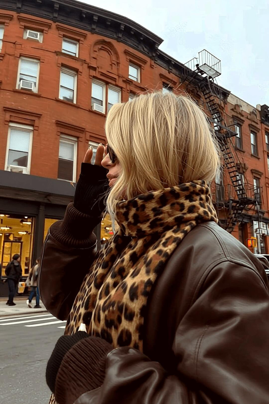 Marais leopard scarf at dusk against a brownstone with golden storefront glow — Amavé winter-weight scarf.