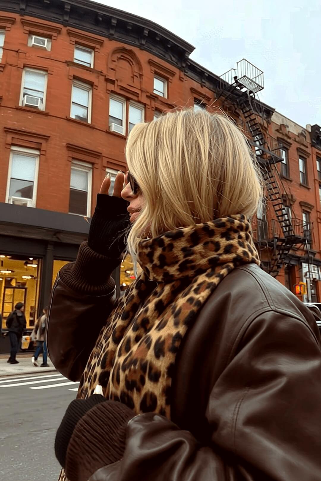 Marais leopard scarf at dusk against a brownstone with golden storefront glow — Amavé winter-weight scarf.