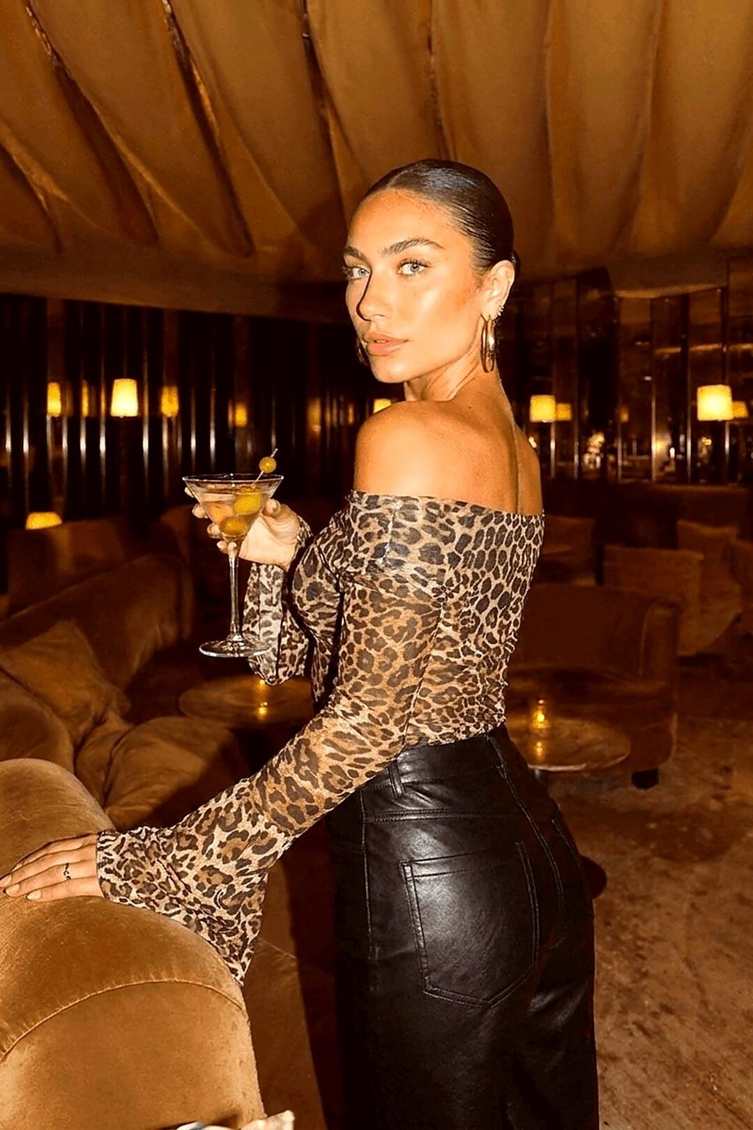 Marseille leopard Bardot top from behind, slicked-back hair and a martini in hand, candlelit interior — Amavé silk Bardot.