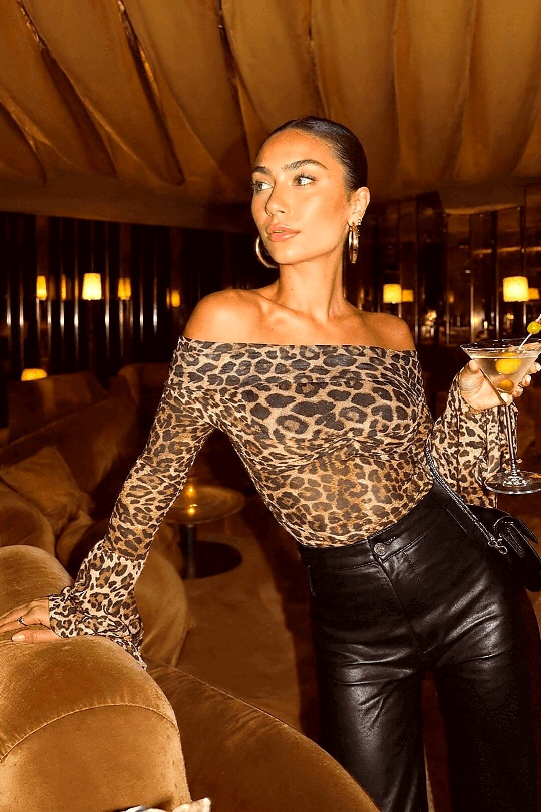 Marseille leopard Bardot top in semi-sheer silk, seated on a camel velvet sofa with a martini, candlelit lounge — Amavé.