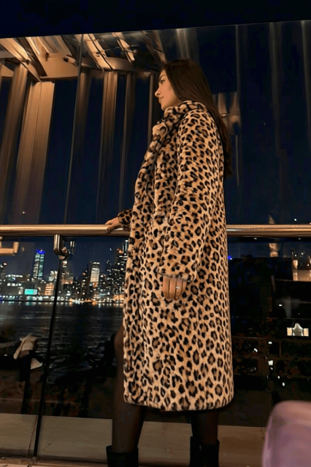 Riviera leopard longline coat on a waterfront terrace, profile facing the illuminated city skyline at night — Amavé after-dark hero.